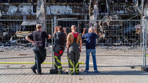 Feuerwehrleute stehen an Zaun vor abgebrannter Fahrzeughalle