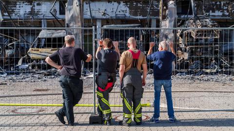 Feuerwehrleute stehen an Zaun vor abgebrannter Fahrzeughalle