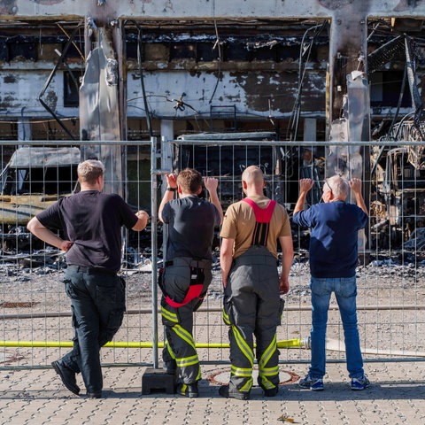 Firefighters stand at a fence in front of a burned-down vehicle hall