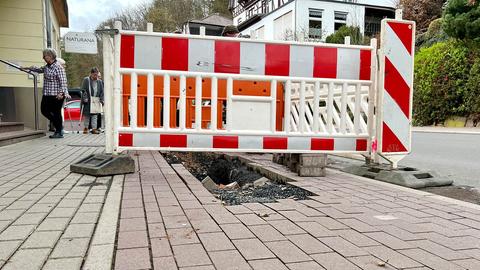 Nahaufnahme eines Bürgersteigs.  In der Mitte befindet sich ein großes Loch, das nur unzureichend von einer rot-weißen Barriere umgeben ist.  Im Hintergrund betreten kleine Menschen mit Gehhilfen das angrenzende Haus. 