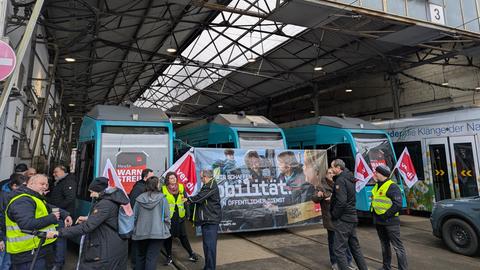 ÖPNV-Mitarbeitende in gelben Verdi-Warnwesten mit Verdi-Fahnen in einem Straßenbahndepot vor Trams