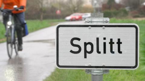 ein Schild mit der Aufschrift "Splitt" steht an einem asphaltierten Feldweg. Im Hintergrund fährt ein Mann auf einem Fahrrad.