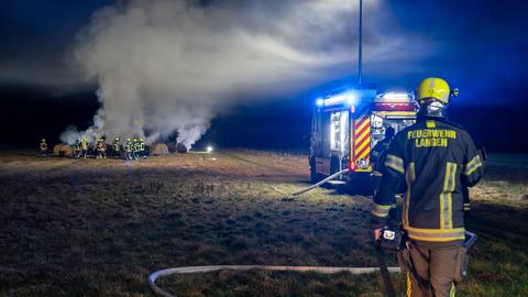 Ein Feuerwehrmann und ein Feuerwehrauto auf einem Feld, im Hintergrund brennende Strohballen, Rauch steigt auf, Löscharbeiten.