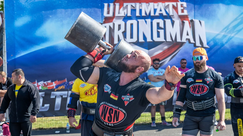 Das Bild zeigt einen Teilnehmer der Strongman Weltmeisterschaft 2024 in Fulda. Er trägt Glatze und einen kurzen Bart und hebt einarmig eine große Hantel hoch. Im Hintergrund sind weitere Teilnehmer sowie ein blaues Banner mit der Aufschrift "Ultimate Strongman" zu sehen. 