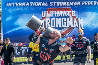 Das Bild zeigt einen Teilnehmer der Strongman Weltmeisterschaft 2024 in Fulda. Er trägt Glatze und einen kurzen Bart und hebt einarmig eine große Hantel hoch. Im Hintergrund sind weitere Teilnehmer sowie ein blaues Banner mit der Aufschrift "Ultimate Strongman" zu sehen. 