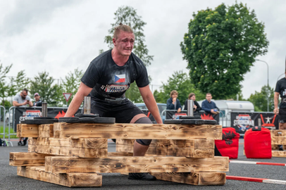 Das Bild zeigt einen Teilnehmer der Strongman Weltmeisterschaft 2024 in Fulda. Ein Mann mit dunkelblonden, zum Zopf gebundenen Haaren und schwarzem T-Shirt eine Konstruktion aus Europaletten, auf denen Gewichte liegen. Er schaut angestrengt.