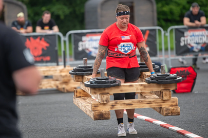 Das Bild zeigt eine Teilnehmerin der Strongman Weltmeisterschaft 2024 in Fulda. Eine Frau mit dunklen, zum Zopf gebundenen Haaren und rotem T-Shirt trägt eine Konstruktion aus Europaletten, auf denen Gewichte liegen.