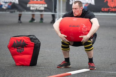 Das Bild zeigt einen Teilnehmer der Strongman Weltmeisterschaft 2024 in Fulda. Ein Mann mit dunkelblonden kurzen Haaren und schwarzem T-Shirt hebt einen roten Sandsack aus den Knien an. Er schaut angestrengt.