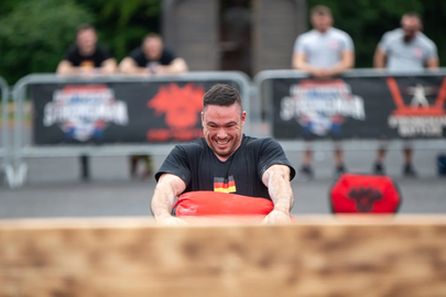 Das Bild zeigt einen Teilnehmer der Strongman Weltmeisterschaft 2024 in Fulda. Ein Mann mit dunklen kurzen Haaren und schwarzem T-Shirt hebt einen roten Sandsack. Er schaut angestrengt.