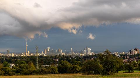 Skyline von Frankfurt aus der Ferne gesehen, im Vordergrund Büsche und Felder, am Himmel türmen sich Regenwolken, durch die Sonnenstrahlen brechen