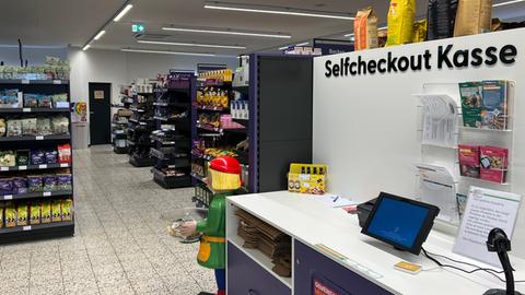 Blick in einen Supermarkt. Im Hintergrund sind Regale voller Waren, im Vordergrund ein Kassensystem, bei dem man selbst kassieren kann.