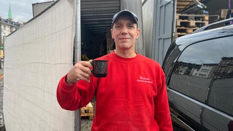 Ein Weihnachtsmarktstand-Betreiber zeigt eine Glühwein-Tasse, die er nun wegen Unbrauchbarkeit zurückgibt. Auf dem Pullover des Mannes steht der Aufdruck "Elsässer Flammkuchen".