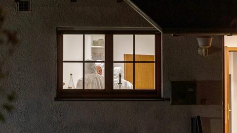 Durch ein Fenster einer im Dunkeln liegenden Fassade sind ein hell erleuchteter Raum mit drei Menschen in weißen Ganzkörperanzügen zu sehen.