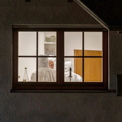 Durch ein Fenster einer im Dunkeln liegenden Fassade sind ein hell erleuchteter Raum mit drei Menschen in weißen Ganzkörperanzügen zu sehen.