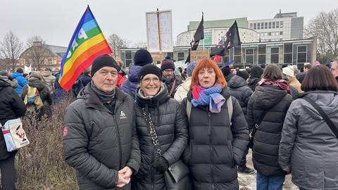 Ein Mann und zwei Frauen vor einer Menschenmenge. Sie tragen dicke schwarze Jacken, der Mann und eine Frau auch Mützen.