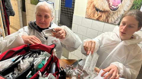 Zwei Personen in weißen Schutzanzügen sortieren medizinische Utensilien. In der Mitte befindet sich eine offene, rote Tasche mit Medikamenten und Spritzen. Auf dem Tisch liegen weitere medizinische Materialien und ein Blatt mit Text. Im Hintergrund ist ein großes Foto eines Hundes zu sehen.