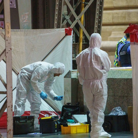 Menschen in weißen Ganzkörperanzügen und blauen Handschuhen untersuchen eine Stelle im Bahnhof.