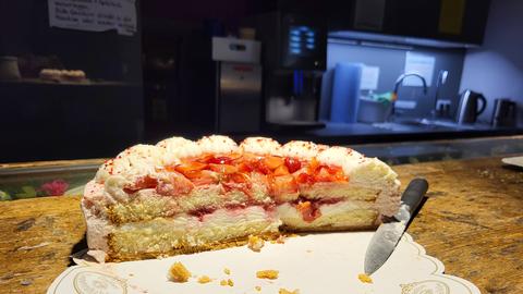 Ein angeschnittener Erdbeerkuchen mit Sahnehaube steht auf einem Holztisch. Neben dem Kuchen liegt ein Messer. Der Kuchen befindet sich auf einer Unterlage mit der Aufschrift "Coppenrath & Wiese". Im Hintergrund ist eine Küche mit beleuchteter Spüle und Wasserkocher zu sehen.