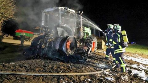 Feuerwehrleute nachts auf einem Feld löschen mit Wasser aus einem Rohr einen Traktor, von dem nur noch Räder und Gerippe zu sehen sind