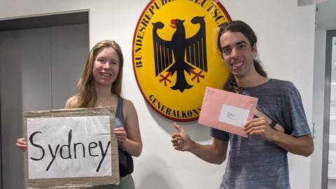 Ein Mann und eine Frau stehen vor einem Schild mit der Aufschrift "Bundesrepublik Deutschland, Generalkonsulat"