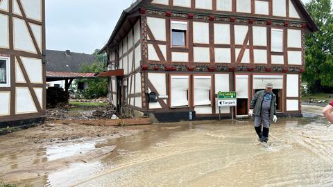 Mit braunem Wasser überflutete Straße, die an einem Fachwerkhaus vorbeiführt. Vor dem Haus steht ein Mann mit Gummistiefeln knöcheltief im Wasser.