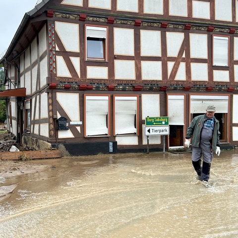 Mit braunem Wasser überflutete Straße, die an einem Fachwerkhaus vorbeiführt. Vor dem Haus steht ein Mann mit Gummistiefeln knöcheltief im Wasser.