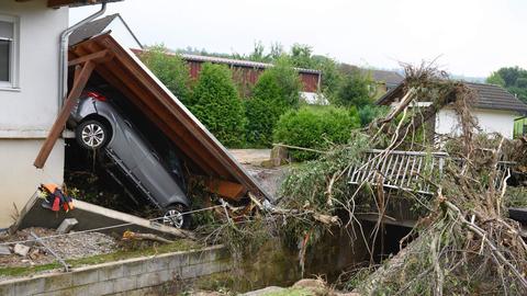 Zerstörte Garage mit einem am Haus hochgedrücktem Auto. Daneben eine kleine zerstörte Brücke mit viel angestautem Geäst und Unrat.