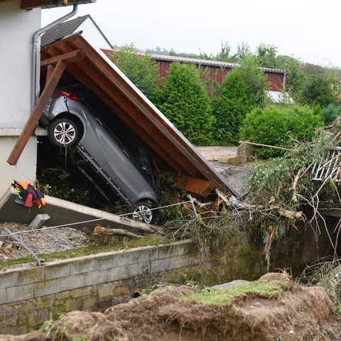 Zerstörte Garage mit einem am Haus hochgedrücktem Auto. Daneben eine kleine zerstörte Brücke mit viel angestautem Geäst und Unrat.