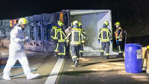 Lastwagen liegt auf Autobahn und blockiert diese, man sieht ihn von unten. Feuerwehrleute und Einsatzkräfte in Schutzanzügen laufen auf der Fahrbahn