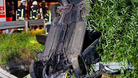 Feuerwehrleute stehen hinter einer Leitplanke. Ein Auto liegt davor falsch herum in einem Graben, es ist von hinten beziehungsweise unten zu sehen, die Planke ist verbogen.