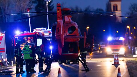 Ein Rettungshubschrauber steht auf einer abendlichen Straße, umgeben von Personen in Einsatzkleidung. Im Hintergrund sind beleuchtete Einsatzfahrzeuge mit Blaulicht zu sehen. Eine Kirche ist im Hintergrund leicht beleuchtet erkennbar. Verkehrsleitkegel stehen auf der Straße.