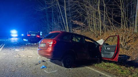 Weinroter Kleinwagen mit offener Beifahrertür halb im Straßengraben, dahinter ein dunkler Pkw auf dem Dach liegend, auf der Straße verteilte Trümmerteile