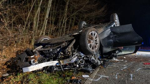 Auf dem Dach im Straßengraben liegendes schwarzes Auto mit geöffneter Beifahrertür, die Front des Autos ist völlig zerstört.