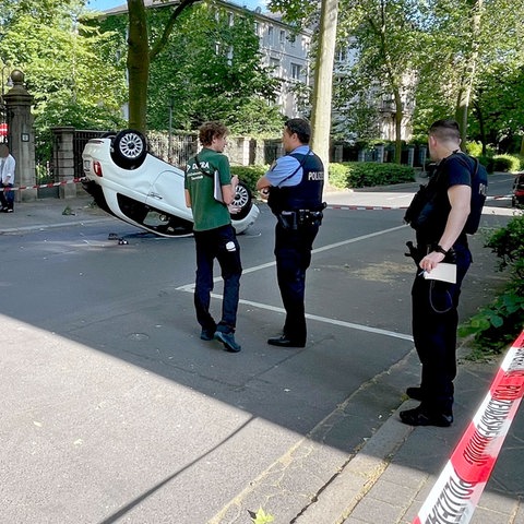 In einer Straße liegt ein Cabrio liegt nach einem Überschlag auf seinem Dach. Polizeikräfte stehen daneben.