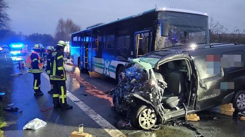 Zerstörter Pkw im Vordergrund, dahinter ein Linienbus mit zerstörter Front, daneben drei Feuerwehrleute