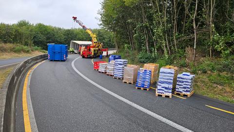 Lebensmittel auf Paletten stehen auf deiner Autobahn vor einem umgekippten Lastwagen, der mit einem Kran wieder aufgerichtet werden soll.