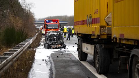 ausgebrannte Überreste eines Smart auf einem Randstreifen der Straße, daneben der gelbe Lkw und rundherum Rettungskräfte