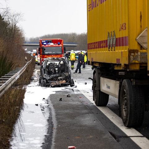 ausgebrannte Überreste eines Smart auf einem Randstreifen der Straße, daneben der gelbe Lkw und rundherum Rettungskräfte