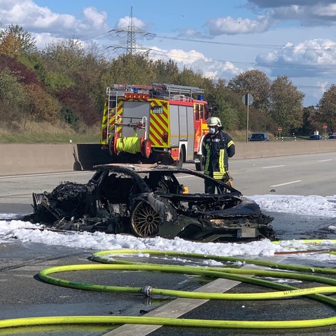 Zwei völlig ausgebrannte Autos liegen auf der Fahrbahn. Sie sind von Löschschaum bedeckt. Feuerwehrleute stehen daneben. 