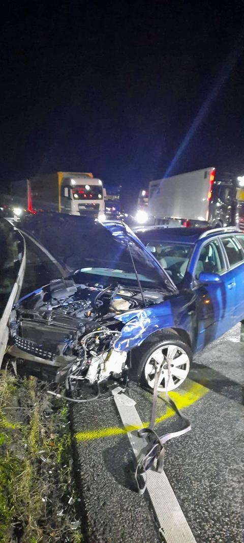 Zerstörter blauer Pkw mit offener Motorhaube, dahinter zwei Laster, Dunkelheit