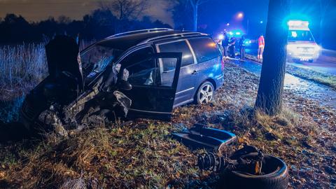 Zerstörter Van neben einem Baum, dahinter eine Landstraße und Rettungskräfte sowie eine Notarztwagen mit Blaulicht, ansonsten Dunkelheit