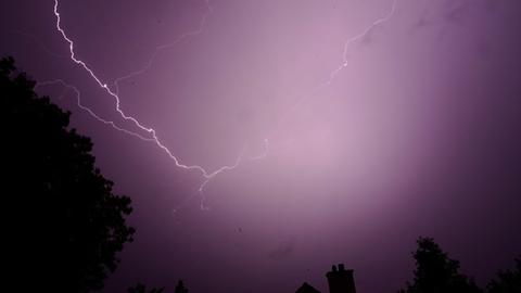 Ein Blitz vor einem violetten Himmel, im Hintergrund stehen Wohnhäuser und Bäume.