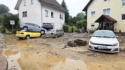 Straße mit viel Schlamm und braunem Wasser, im Hintergrund zwei Wohnhäuser. In der Mitte vier Autos, die chaotisch überneinander liegen und eins das daneben steht.