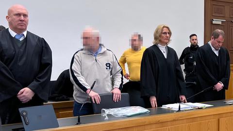 In einem Gerichtssaal stehen zwei Angeklagte mit verpixelten Gesichtern. Um sie herum stehen diverse Rechtsanwälte und Polizisten.