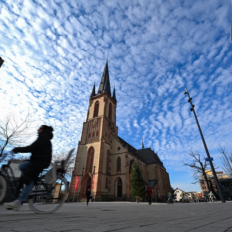 Platz mit einer Kirche, eine Radfahrerin ist verschwommen im Vordergrund zu erkennen.