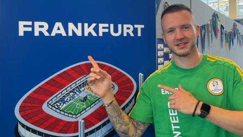 Moritz Kolb - Volunteer -  in grünem Shirt zeigt lächelnd auf einen EURO 2024 Aufsteller