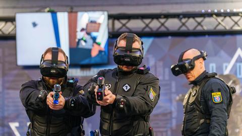 Drei Polizisten stehen in Uniform mit VR-Brille vor einem Monitor, in der Hand halten sie Spielzeug-Pistolen.