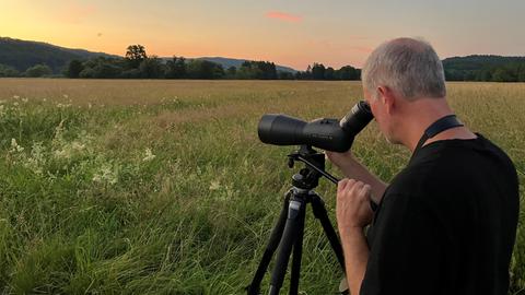 Ein Mann mit großem Fernglas steht vor einer Wiese 