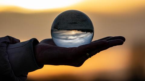 Ein Frau hält eine Glaskugel in der Hand, in der sich die Umgebung und der Sonnenuntergang spiegeln. 