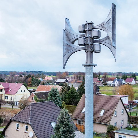 Nahaufnahme einer Sirene auf einem Dach. Darunter/hinter kleinen Häusern eines Dorfes und der umgebenden Landschaft sowie dem Himmel.